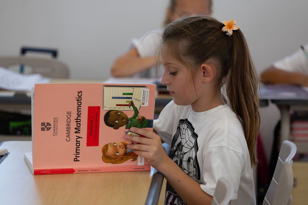 A pupil holding a Cambridge Primary Mathematics workbook in a Golden Oak classroom.
